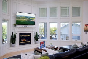 Ein Wohnzimmer mit Kamin, einem darüber montierten Fernseher und einem großen Fenster mit Blick auf ein Haus.