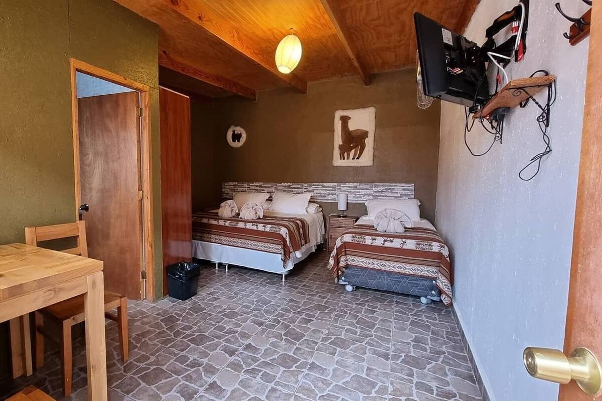 Quarto com duas camas, uma escrivaninha de madeira, uma cadeira, piso de ladrilho e teto de madeira.