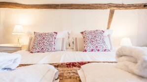 A hotel room with a wooden ceiling, two beds with floral pillows, a nightstand with a lamp, and towels on the floor. - Angel Cottage, Wenhaston. Nr Southwold and Minsmere (Suffolk)
