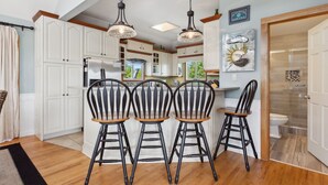 A kitchen with white cabinets, a wooden dining table with four chairs, pendant lights, and a view of the outdoors. - 2135 Pirates Perch * 11 Min Walk to Beach (Duck)