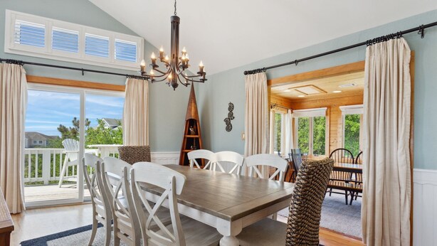 A dining area with a wooden table and white chairs, a chandelier, and a view of the outdoors. - 2135 Pirates Perch * 11 Min Walk to Beach (Duck)
