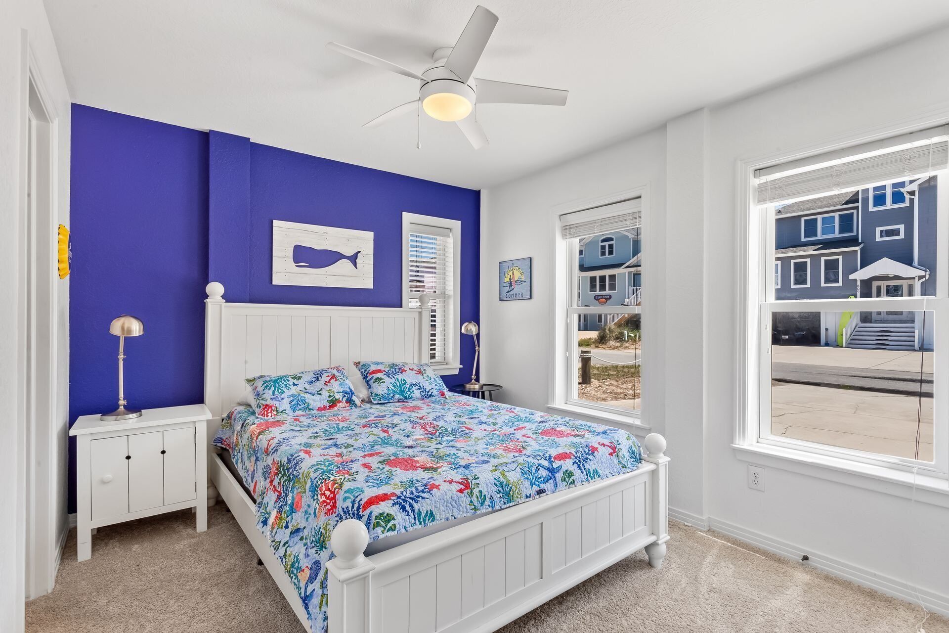 A bedroom with a bed, a nightstand, a ceiling fan, a window with a view of buildings, and a wall with a whale painting.