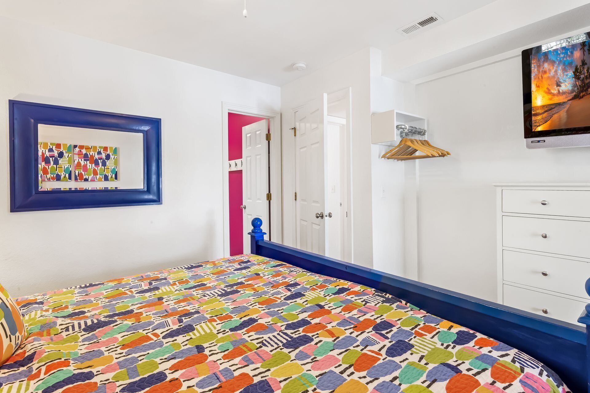 A bedroom with a colorful bedspread, a framed artwork, a wardrobe, and a television on the wall.