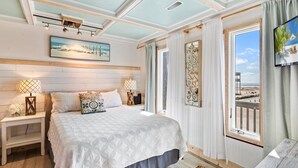 Une chambre avec un grand lit, deux tables de chevet avec des lampes, un tableau représentant une scène de plage et une vue sur la plage par la fenêtre.