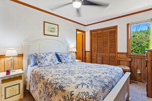 A bedroom with a bed, nightstands, a ceiling fan, wooden wardrobe, and a window with a view of greenery.