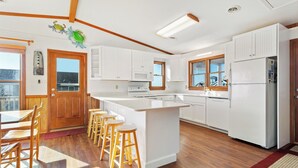 Une cuisine moderne avec des meubles blancs, un îlot central doté de tabourets et une vue sur l’extérieur grâce à de grandes fenêtres.