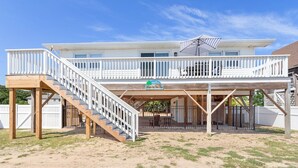 Ein zweistöckiges Strandhaus mit einer Holzterrasse, einer überdachten Veranda und einer Treppe, die zum oberen Stockwerk führt.