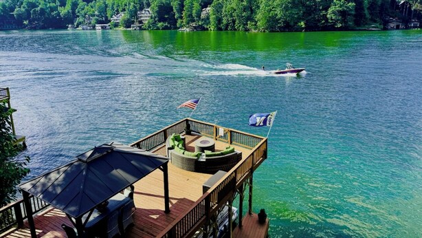 A wooden deck with outdoor seating and a flag, overlooking a lake with a boat in the distance and mountains in the background.