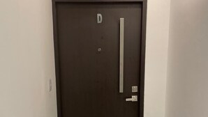 Una puerta de madera oscura con la letra 'D' en la parte superior, un picaporte vertical y una pequeña rejilla rectangular en la esquina inferior izquierda.