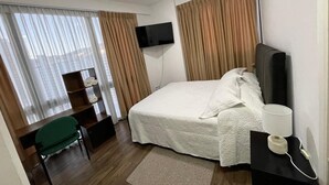 Habitación de hotel con cama, escritorio, silla, televisor y ventana con cortinas.