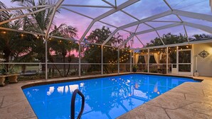 An outdoor swimming pool with a clear blue water surface, surrounded by a covered area with string lights and palm trees. - Canalside 4BR villa with pool, spa, dock, and Gulf access. (Cape Coral)