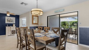 A dining area with a wooden table set for six, surrounded by upholstered chairs. A ceiling fan and pendant light are visible above the table. - Canalside 4BR villa with pool, spa, dock, and Gulf access. (Cape Coral)