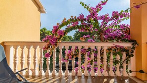 Un balcón con barandilla blanca, flores de buganvilla rosadas y un cielo azul despejado al fondo.