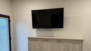 Una habitación con un televisor de pantalla plana montado en la pared, un mueble de madera debajo y una puerta con persianas.