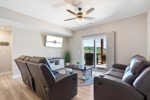 A modern living room with a ceiling fan, a flat-screen TV mounted on the wall, a sliding glass door leading to an outdoor area, and two leather sofas.