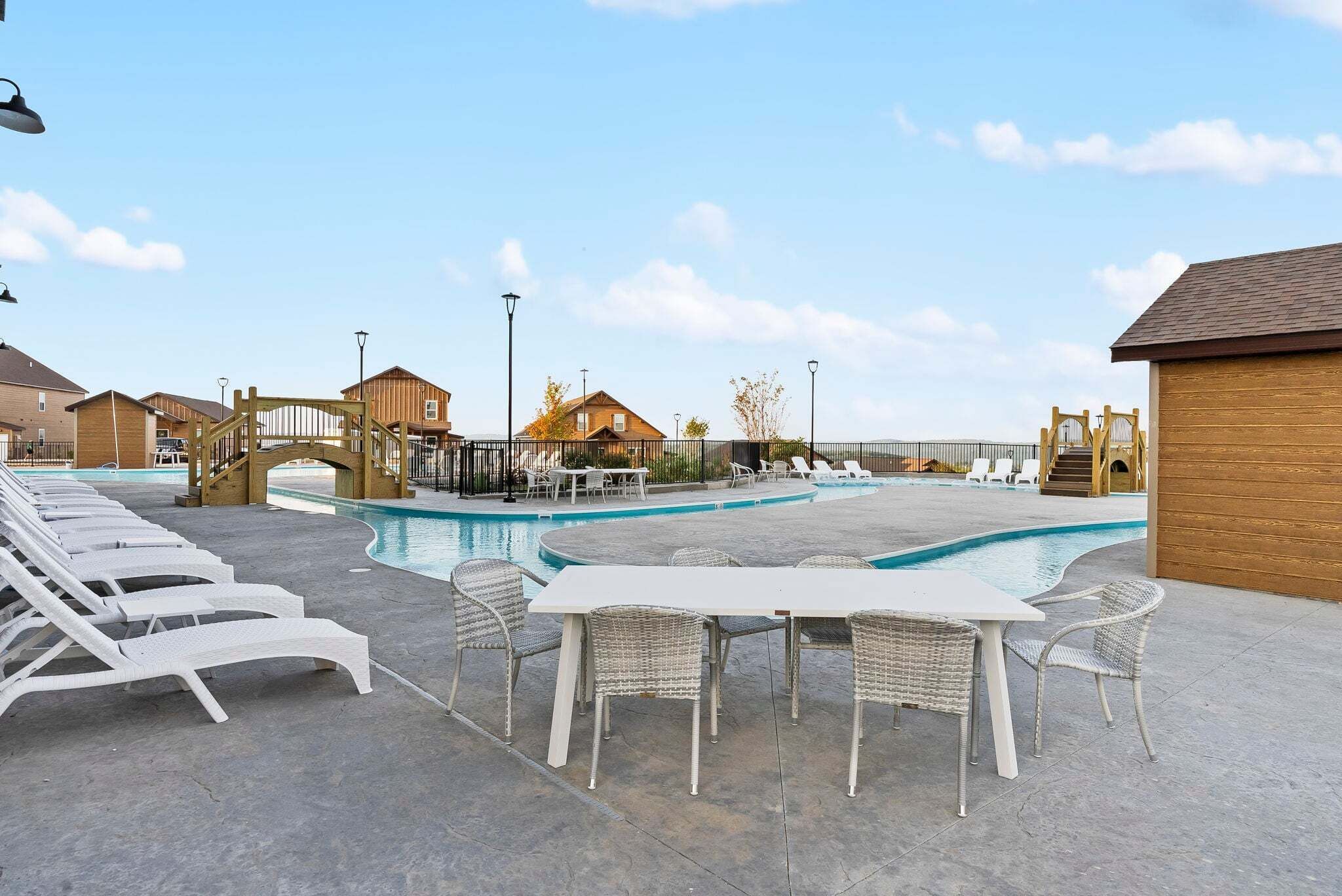 A pool area with lounge chairs and a table, wooden buildings, and a bridge.