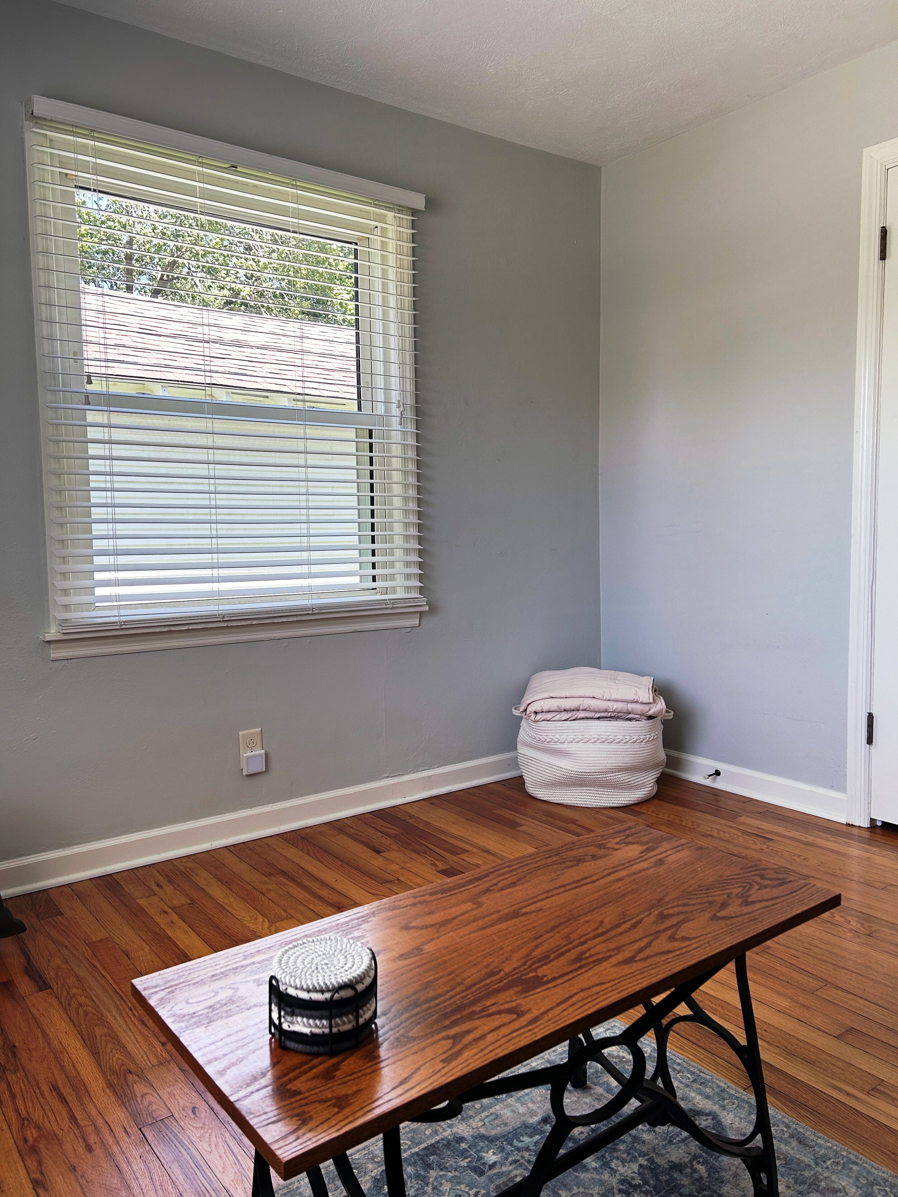 Una habitación con piso de madera, una ventana con persianas, una puerta blanca y una pequeña mesa redonda con una cesta tejida.