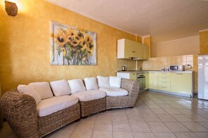 A living room with a sofa, a painting of sunflowers, a kitchen with yellow cabinets, and a tiled floor. - GuestHost - Porto Cervo Two-Levels Apt. W/ Balconies & Parking (Porto Cervo)