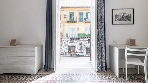 Una habitación con vistas a un balcón, un escritorio blanco, una silla, un piso estampado y un cuadro en la pared.