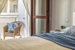 Ein Schlafzimmer mit einem Bett, einem Korbsessel, einem Nachttisch mit einem Buch und Blick durch eine Glastür.