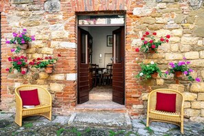 A stone building with a wooden door, two wicker chairs with red cushions, and potted flowers on the walls.