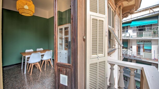 A room with a dining table and chairs, a wooden door, a balcony with a railing, and a building with 'Atriums Apartments' sign.