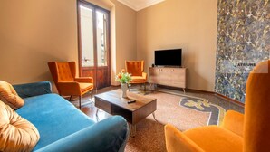 A living room with a blue sofa, orange armchairs, a coffee table with a vase, a flat-screen TV, and a patterned wall.