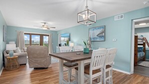 Un coin repas avec une table en bois et des chaises blanches, un ventilateur de plafond et une vue sur l’extérieur grâce à de grandes fenêtres.