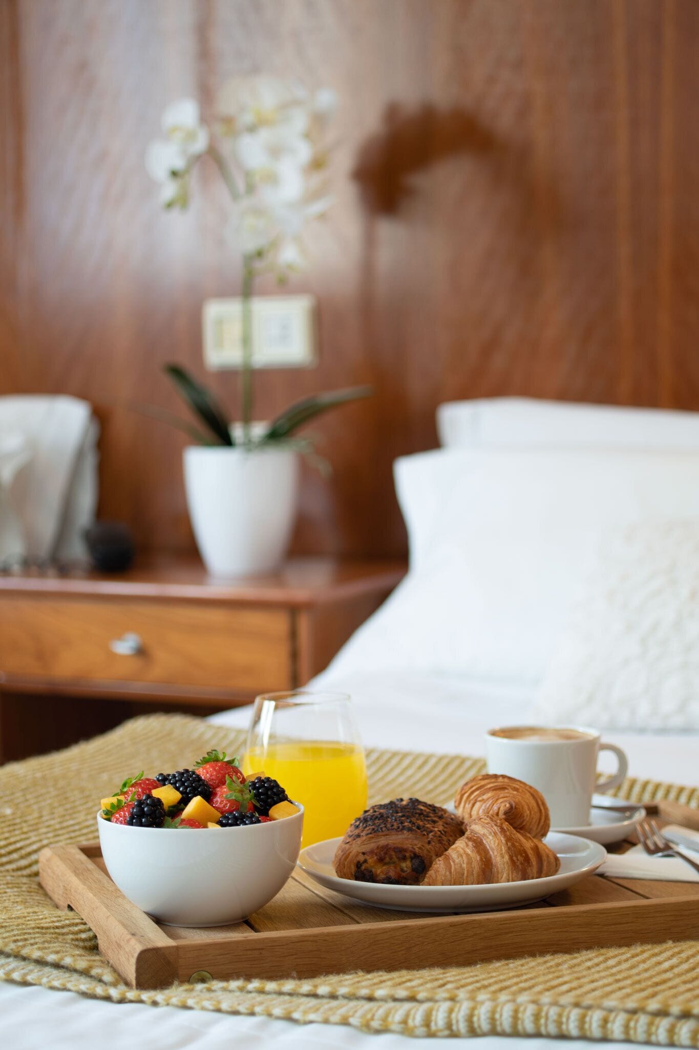 A breakfast tray with a bowl of fruit, croissants, a glass of orange juice, and a cup of coffee on a woven mat.