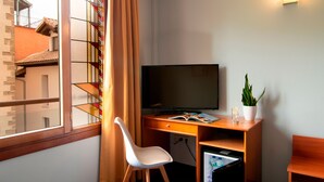Une chambre d’hôtel avec un bureau en bois, une télévision, une chaise blanche, un petit réfrigérateur, une plante en pot et une fenêtre avec des rideaux.