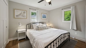 Une chambre à coucher comprenant un lit, des tables de chevet, un ventilateur de plafond et deux fenêtres avec des rideaux.