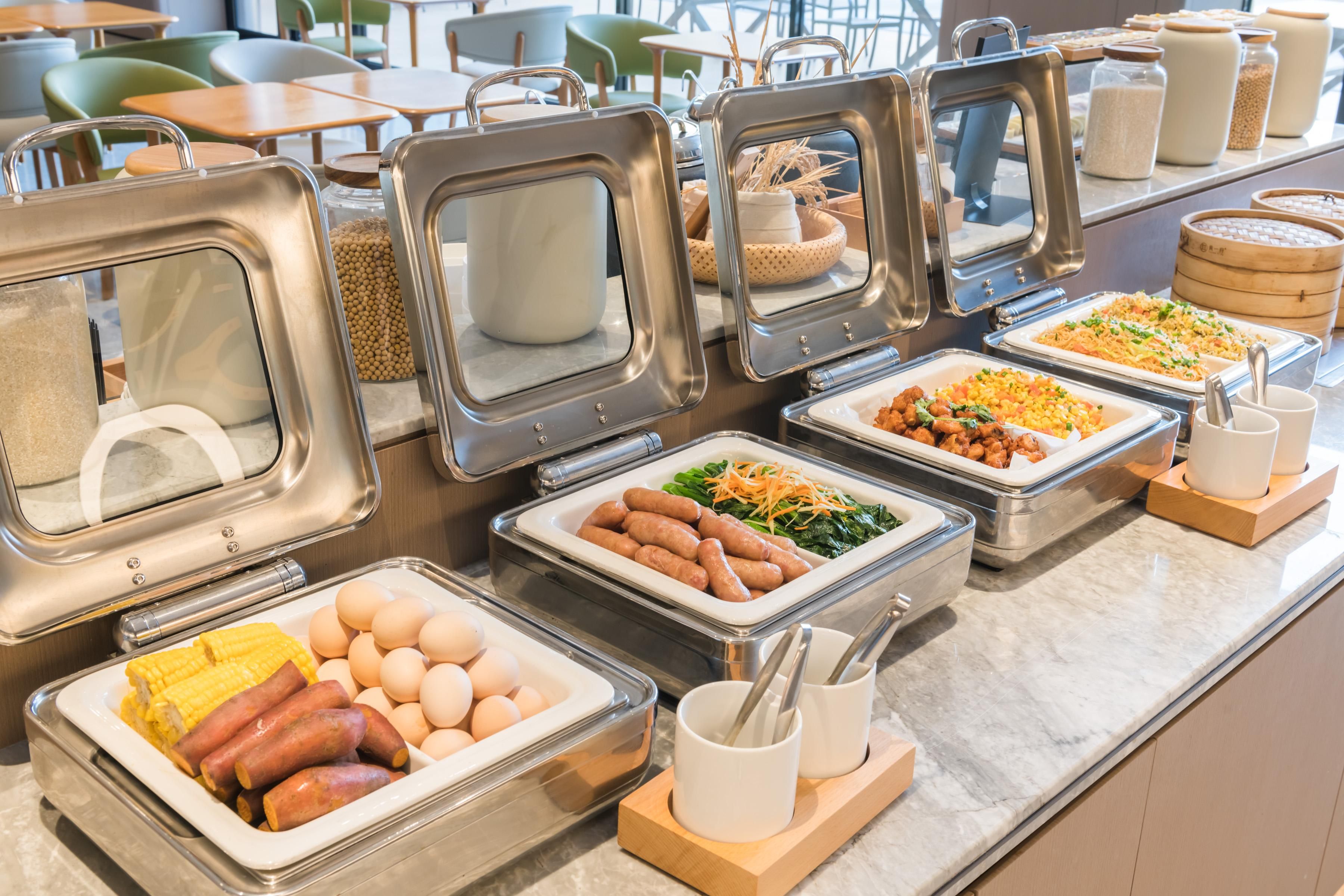Un bufé con diversos alimentos en recipientes metálicos, como salchichas, huevos, maíz y ensaladas.