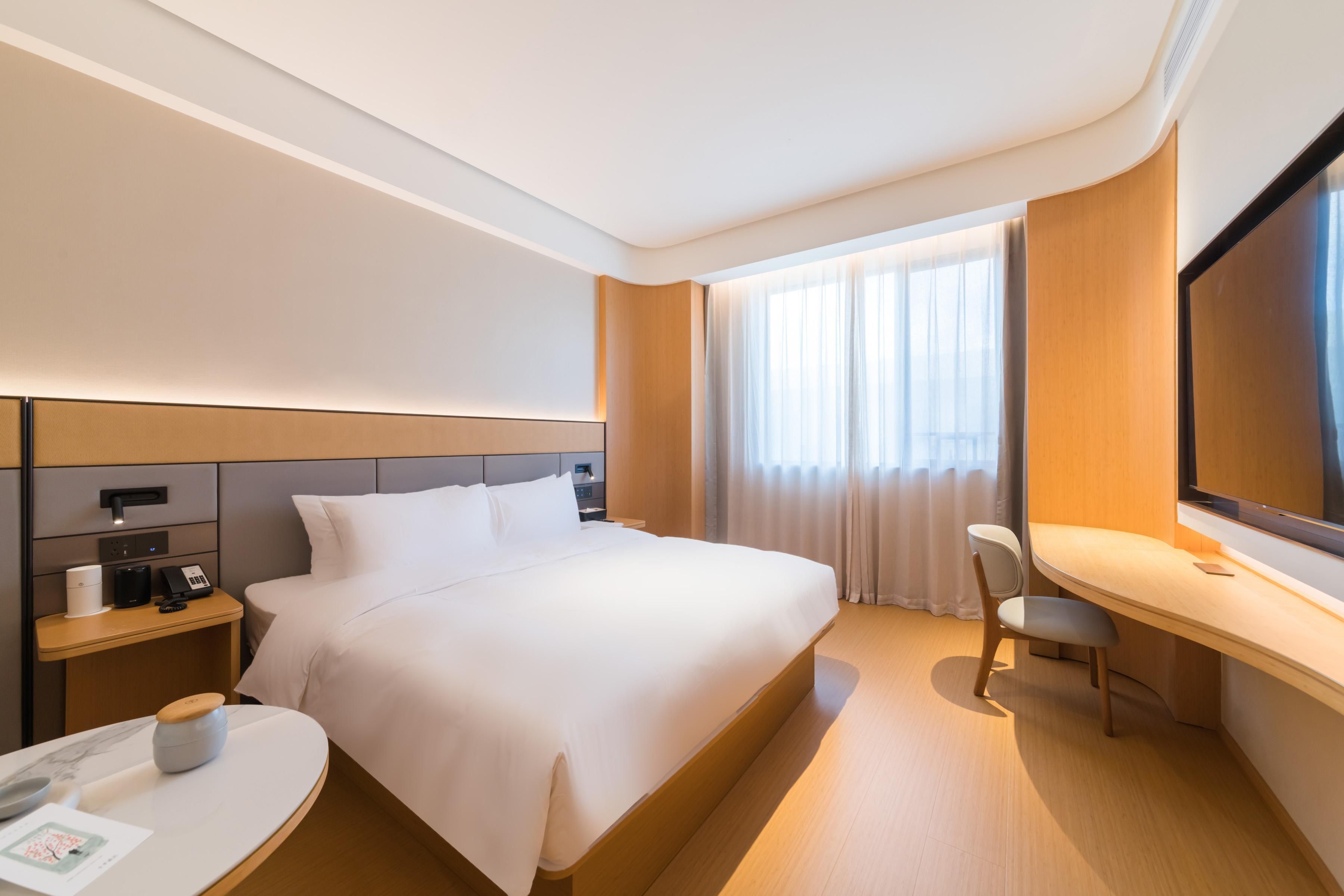 A hotel room with a large bed, bedside tables, a desk, a chair, a flat-screen TV, and a window with curtains.