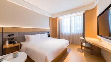 A hotel room with a large bed, bedside tables, a desk, a chair, a flat-screen TV, and a window with curtains.