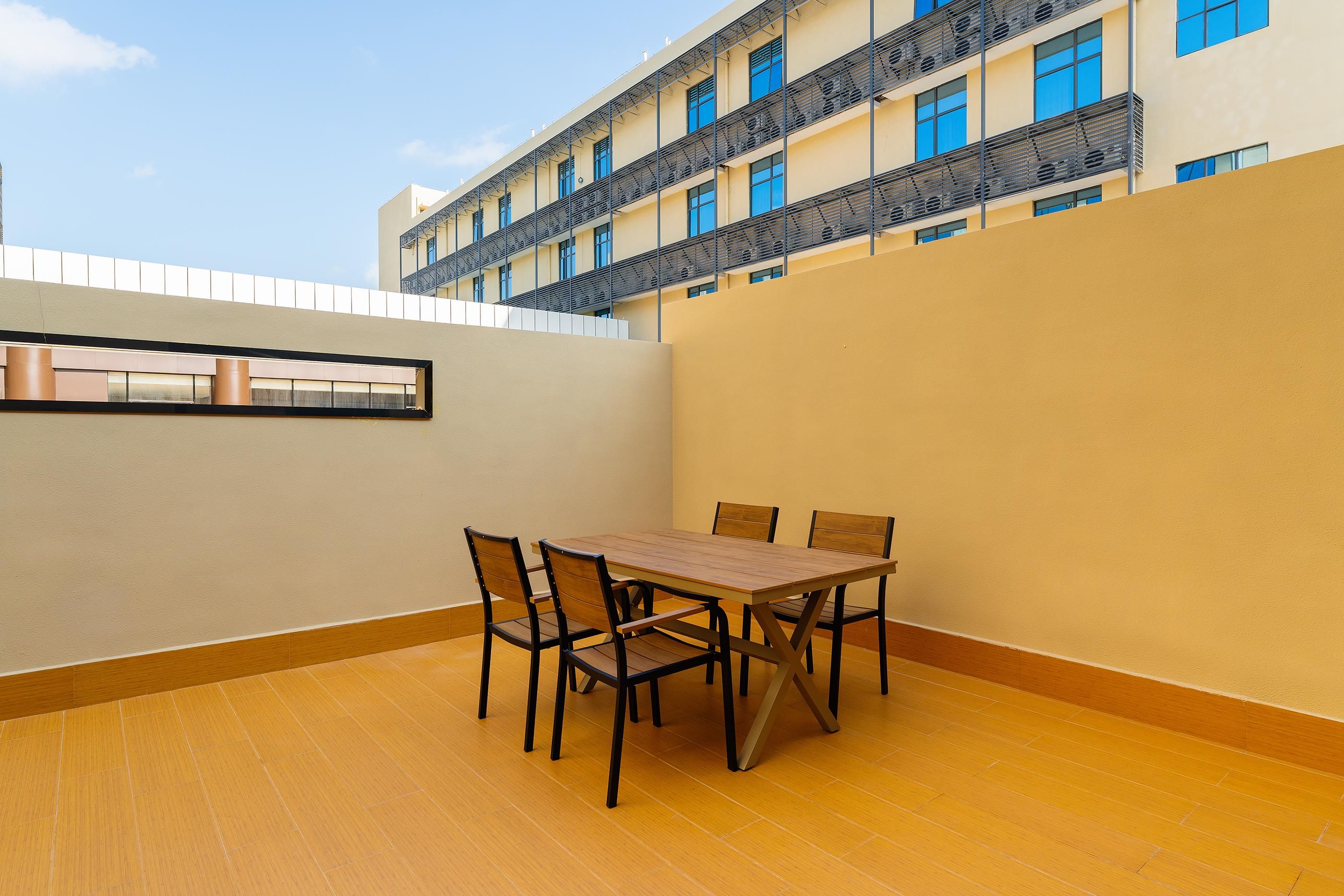 A dining area with a table and chairs, a view of a building with balconies, and a clear sky.