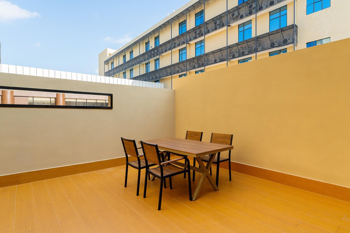 A dining area with a table and chairs, a view of a building with balconies, and a clear sky.