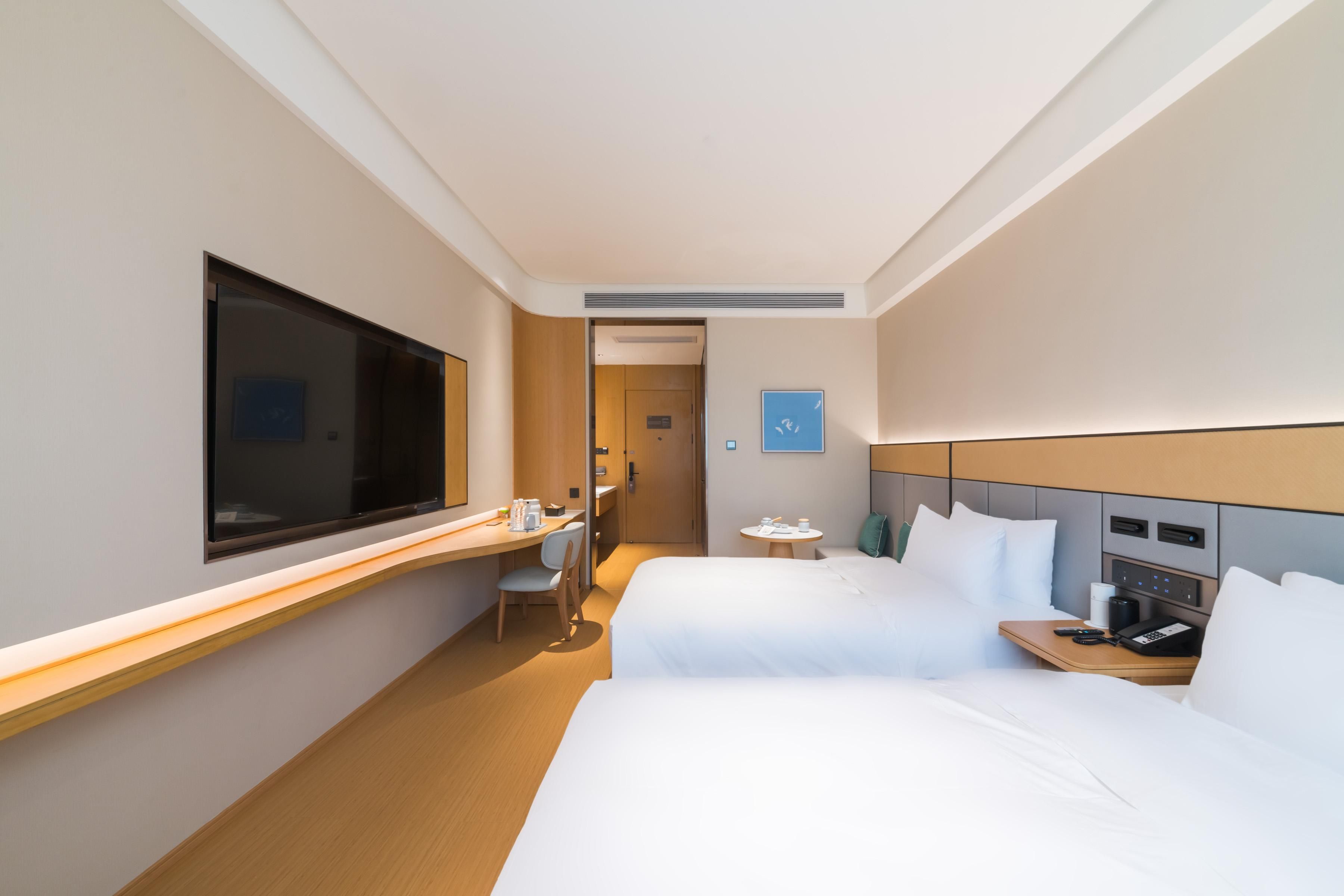 A hotel room with two beds, a desk, a chair, a TV, and a small table with a phone and a cup.