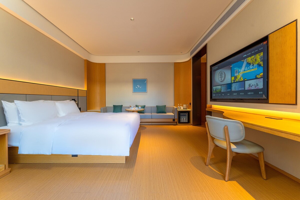 A modern hotel room with a large bed, a desk with a chair, a television, and a small table with a lamp.