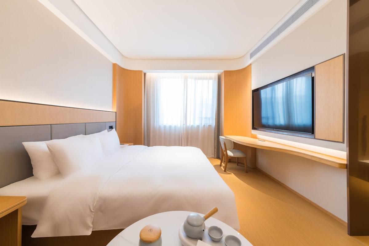 Hotellihuone, jossa on suuri sänky, työpöytä tuolilla, televisio sekä pieni pöytä, jonka päällä on teekannu ja kuppeja.
