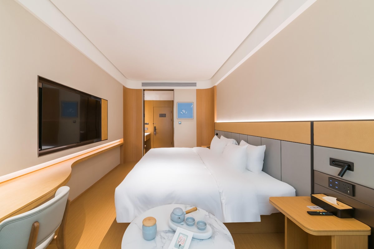 A modern hotel room with a large bed, bedside table, and a desk with a chair.