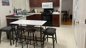 A compact kitchen with a white countertop, dark wooden cabinets, a white microwave, and a black stove. There is a small dining area with a white table and four stools. - CALM Guest House (Auburn)