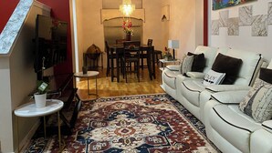 A living room with a white sofa, a dining table with chairs, a TV mounted on the wall, and a red accent wall. - CALM Guest House (Auburn)