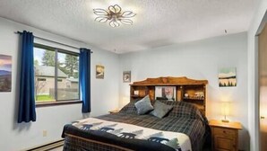 A bedroom with a bed, a nightstand, a lamp, a window with curtains, and a ceiling light fixture. - Cedar Trees Retreat (Sandpoint)