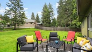 An outdoor patio with a table and four chairs, surrounded by a wooden deck and a grassy yard with trees. - Cedar Trees Retreat (Sandpoint)