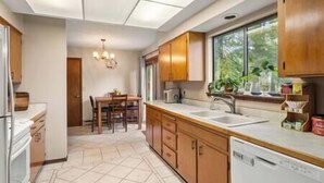 A kitchen with wooden cabinets, a dishwasher, a window with plants, and a dining area with chairs and a table. - Cedar Trees Retreat (Sandpoint)