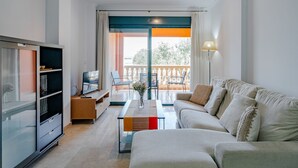 A modern living room with a sofa, coffee table, TV, and bookshelf. There is a balcony with a view of greenery.