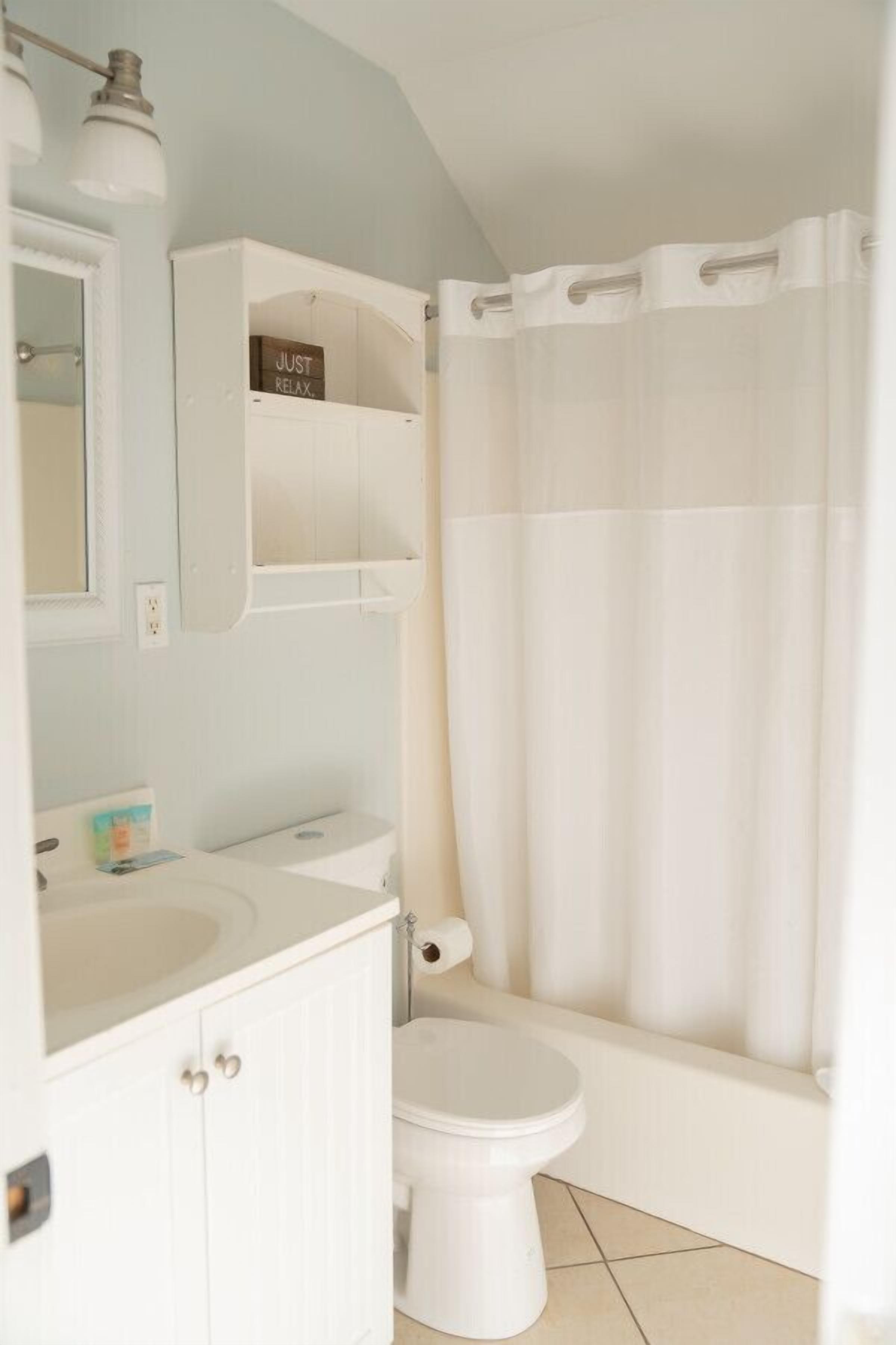Un baño con lavamanos, inodoro y bañera blancos. Hay un estante sobre el lavamanos con un letrero que dice “JUST RELAX.”