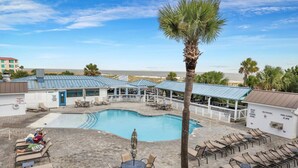 A pool area with lounge chairs, a palm tree, and a building with a blue roof. - Beachfront Condo w Ocean Views and Pool (Tybee Island)