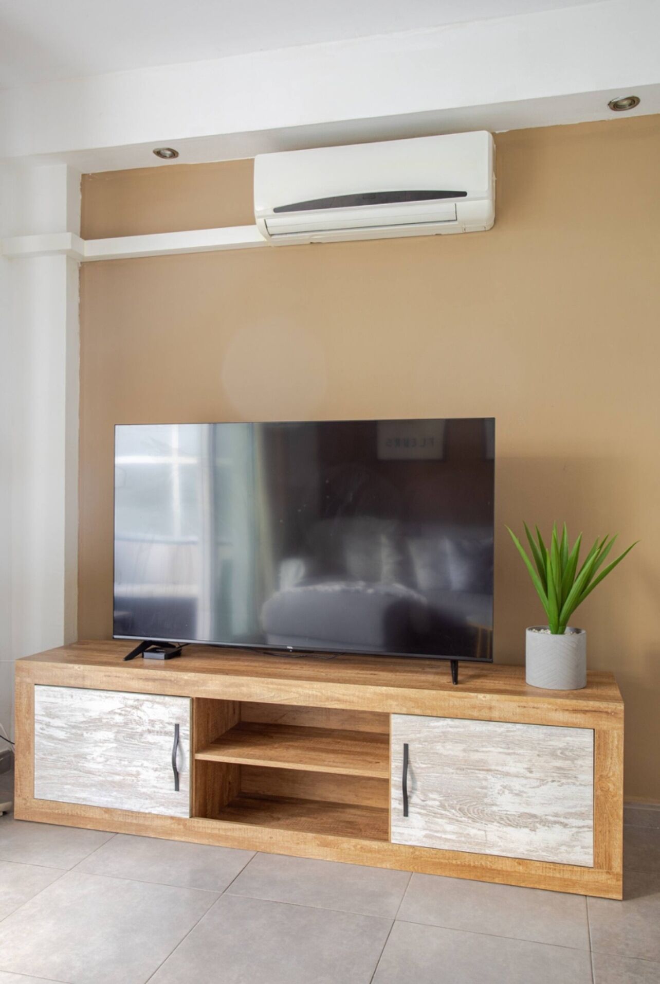 A modern living room with a flat-screen TV on a wooden TV stand with drawers, a small potted plant, and an air conditioning unit mounted on the wall.