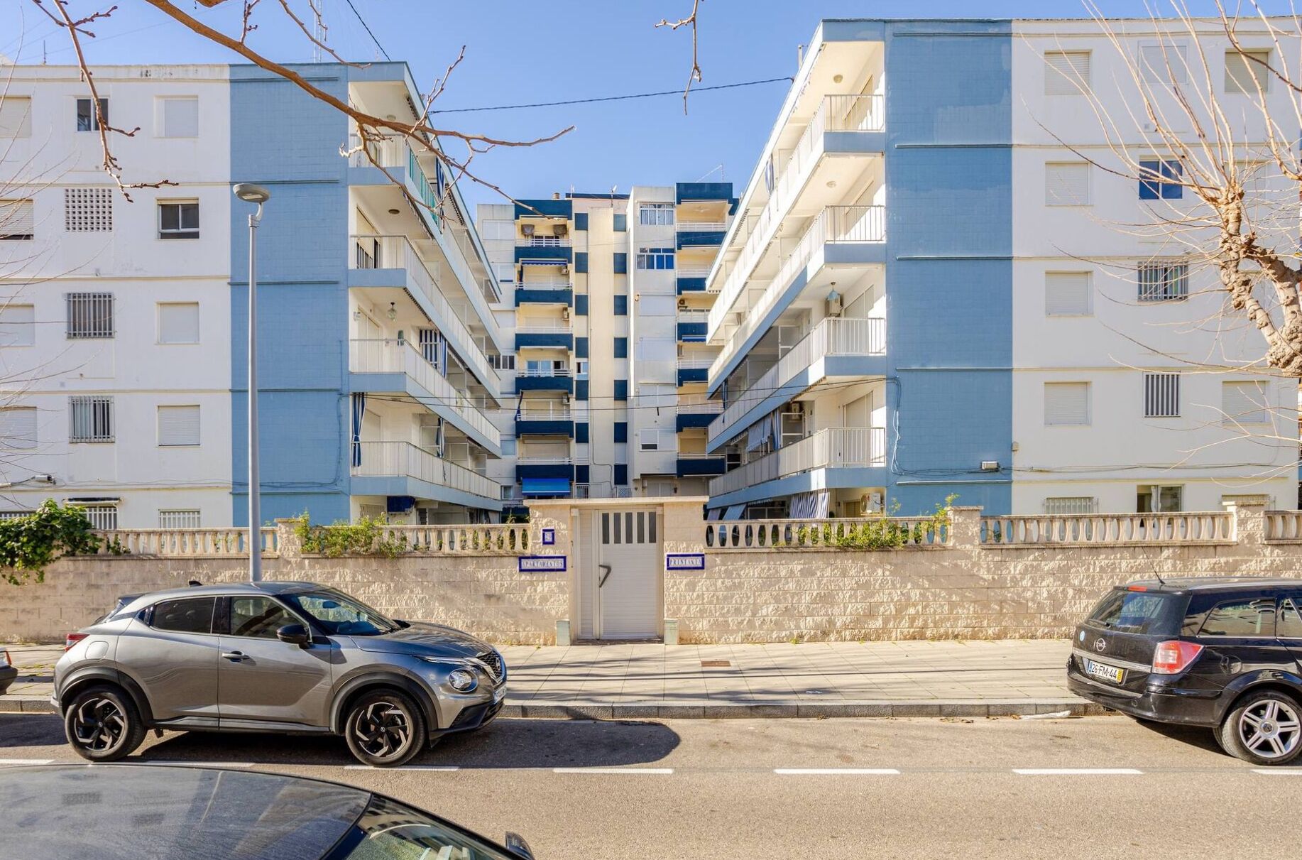 A multi-story residential building with balconies, a gated entrance, and a stone wall.